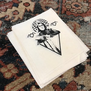 Könnte beinhalten: Ein Stapel weißer Stoffquadrate mit einem schwarz-weißen Grafikdruck. Der Druck zeigt eine Person mit Brille und einem gemusterten Kopfschmuck, umgeben von geometrischen Formen. Der Stoff liegt auf einem gemusterten Teppich.