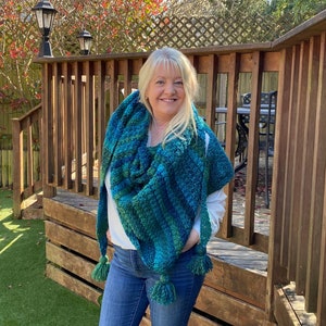 May include: A woman wearing a teal and green striped crocheted shawl with tassels. The shawl is draped around her shoulders and she is standing in front of a wooden deck.