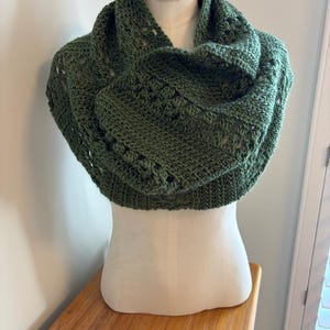 May include: A green crocheted cowl scarf displayed on a mannequin. The scarf is a deep olive green color and features a textured, open-weave design. The cowl is draped around the mannequin's neck and shoulders, showcasing its shape and style.