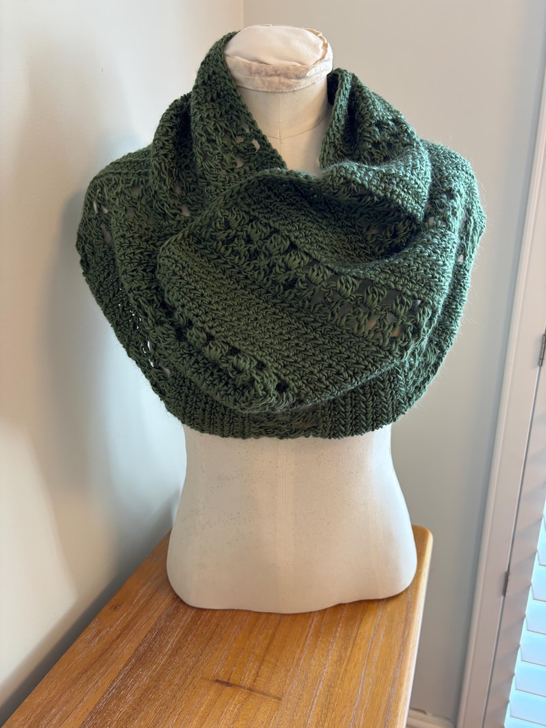 May include: A green crocheted cowl scarf displayed on a mannequin. The scarf is a deep olive green color and features a textured, open-weave design. The cowl is draped around the mannequin's neck and shoulders, showcasing its shape and style.