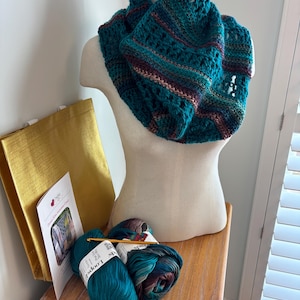 May include: A teal and burgundy crocheted cowl on a mannequin. The cowl features an open, lacy pattern. Two skeins of teal yarn and a crochet hook are next to the cowl. A gold tote bag and a brochure are also in the image.