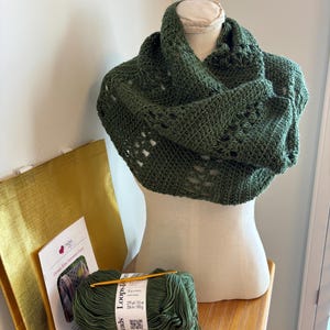May include: A dark green crocheted cowl displayed on a mannequin. The cowl has an openwork design. Beside it are two skeins of Loops & Threads yarn, a crochet hook, and a pattern titled "Ocean Kiss Autumn Cowl". A gold tote bag is also visible.
