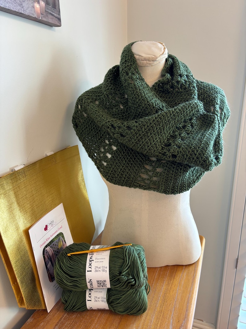 May include: A dark green crocheted cowl displayed on a mannequin. The cowl has an openwork design. Beside it are two skeins of Loops & Threads yarn, a crochet hook, and a pattern titled "Ocean Kiss Autumn Cowl". A gold tote bag is also visible.