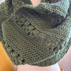May include: A handmade, olive green crochet cowl scarf. The scarf features a textured stitch pattern with openwork details. The cowl is displayed on a mannequin, showcasing its circular design and cozy appearance. The yarn appears soft and warm.