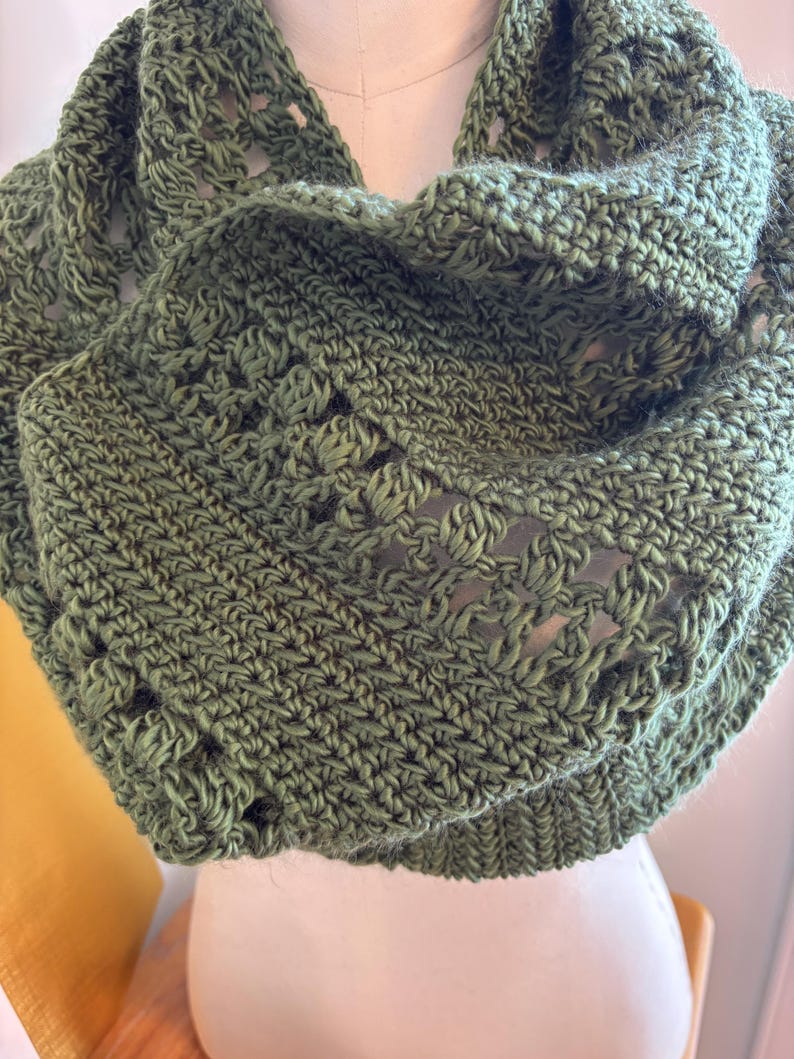 May include: A handmade, olive green crochet cowl scarf. The scarf features a textured stitch pattern with openwork details. The cowl is displayed on a mannequin, showcasing its circular design and cozy appearance. The yarn appears soft and warm.