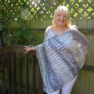 May include: A woman wearing a crocheted poncho with a gray, white, and beige color pattern. The poncho has a loose, open weave and a slightly flared shape.
