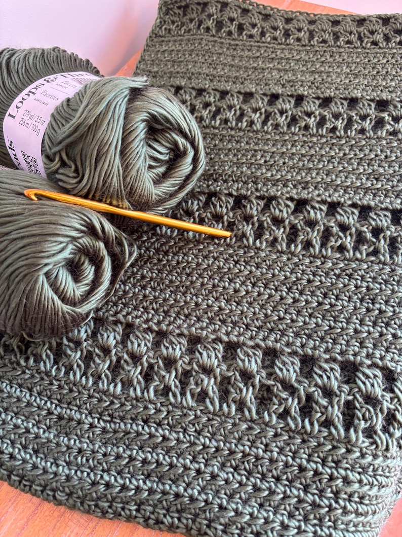 May include: Close-up of olive green crochet yarn and a crochet hook next to a section of a crocheted item. The yarn label reads "Loops & Threads Facettes". The crocheted item has a textured pattern.