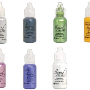 May include: A collection of seven bottles of Liquid Pearls dimensional, pearlescent paint in various colors, including silver, slate, succulent, sunflower, taffy, violet, and white opal. Each bottle has a white dropper top and a label with the product name.