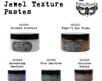 May include: Winter 2022 release of Jewel Texture Pastes by Finnabair.  Four jars of textured paint in various colors: crushed crystals, tiger's eye stone, enchanting amethyst, and true emeralds. Each jar is labeled with the product name and a unique product number.