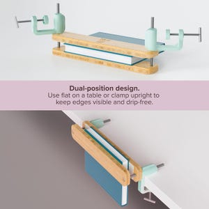 Può includere: Un fermalibri in bambù con morsetti verde menta, che tiene un libro con una copertina verde acqua. Il testo "Dual-position design. Use flat on a table or clamp upright to keep edges visible and drip-free." è visibile.