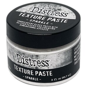 Puede incluir: Un pequeño tarro blanco de pasta de textura Distress Ranger con tapa negra. El tarro está etiquetado con "Distress Texture Paste - Sparkle - 3 fl oz (88.7 ml)".