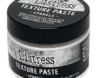 Puede incluir: Un pequeño tarro blanco de pasta de textura Distress Ranger con tapa negra. El tarro está etiquetado con "Distress Texture Paste - Sparkle - 3 fl oz (88.7 ml)".