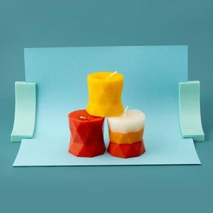 May include: Three pillar candles, one yellow, one white with orange stripes, and one red with a geometric pattern. The candles are arranged in a pyramid shape on a blue background.