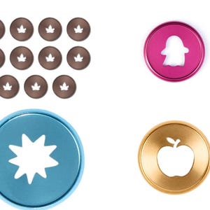 May include: A collection of circular metal discs including eleven small brown discs with maple leaf cutouts, one medium pink disc with a ghost cutout, one large blue disc with a star cutout, and one medium gold disc with an apple cutout.