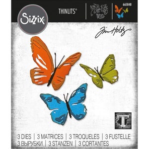 Puede incluir: Tres formas de mariposa troqueladas en naranja, azul y verde. Las troqueles son parte del juego de troqueles Sizzix Thinlits de Jim Holtz.