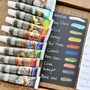 May include: A set of 12 metallic wax tubes in various colours, including Black Gold, Midnight, Coral, Moss Green, Cobalt, Red Flame, Honey, Acid, Sea Glass, and Celestial. The tubes are arranged in a row with the names of the colours written on a black surface.