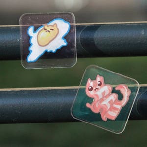 Two square semi-transparent magnets. They have drawings of cats in the shape of food, a friend egg and a strip of bacon, and the charm around them is seethrough. They are photographed stuck on metal railings.