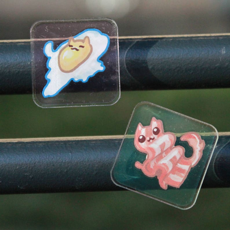 Two square semi-transparent magnets. They have drawings of cats in the shape of food, a friend egg and a strip of bacon, and the charm around them is seethrough. They are photographed stuck on metal railings.