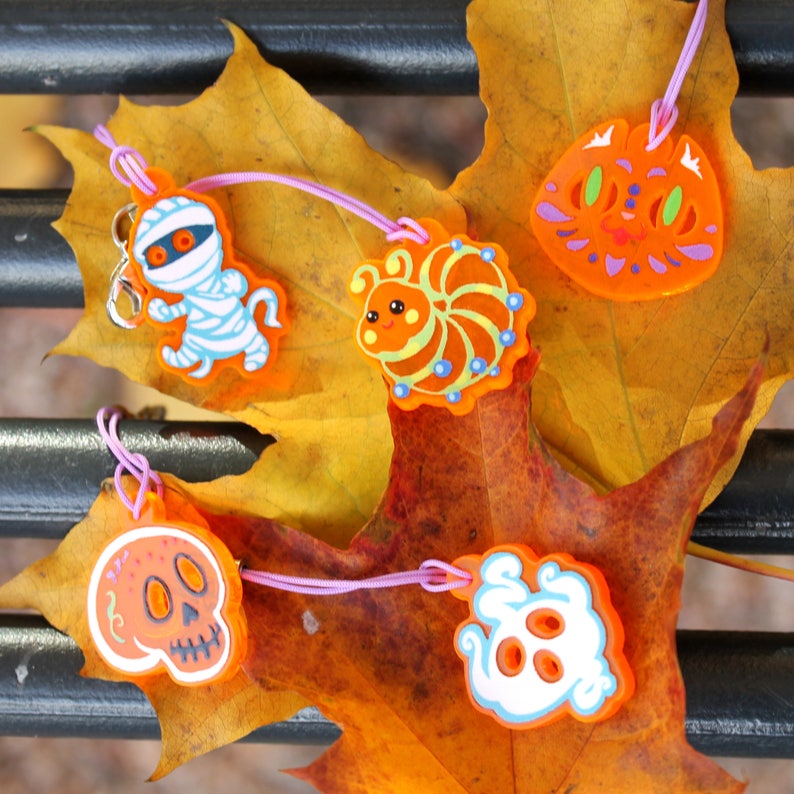 Five orange acrylic phone charm charms photographed in daylight against a yellow maple leaf. There is the mummy, a caterpillar, a cat head, a skull with decorations, and a ghost.