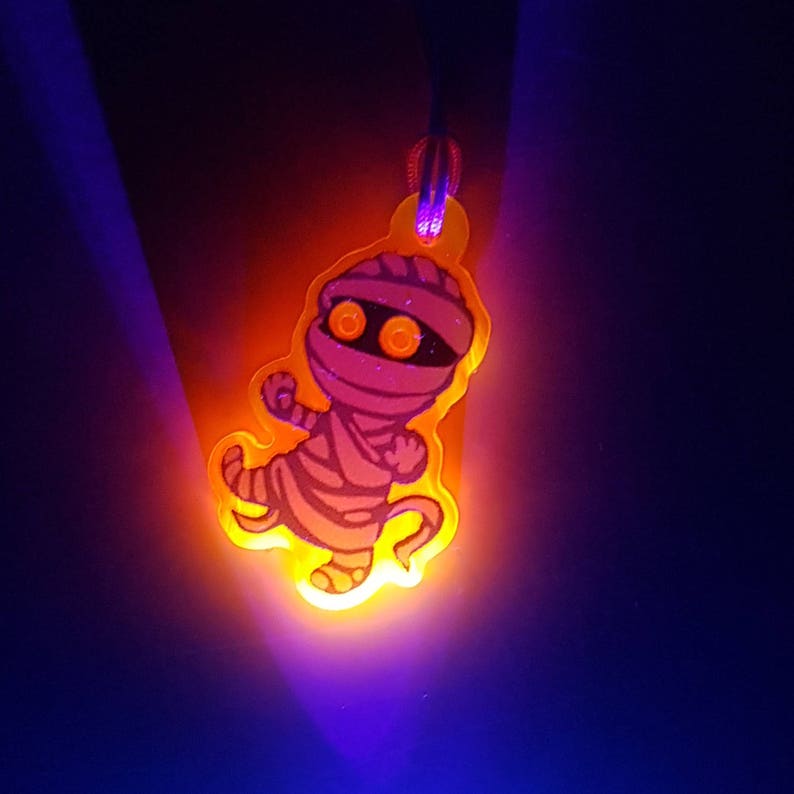 Orange acrylic charm in UV light, the mostly white image of the cartoony mummy looks purple and pink, and the charm has a glowy orange aura around it.