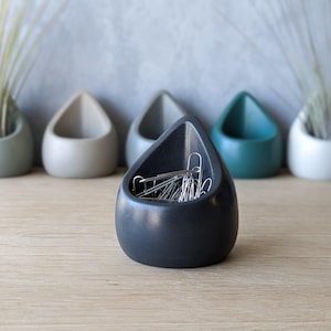 May include: A black ceramic desk organizer shaped like a teardrop, filled with silver paper clips.