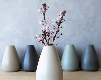 May include: A collection of five teardrop-shaped ceramic vases in varying neutral colors, with a sprig of pink blossoms in the center vase. The vases are arranged in a row on a wooden surface against a gray background.