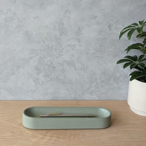 May include: A light green oval-shaped concrete tray with a silver pen inside. The tray is sitting on a wooden surface.