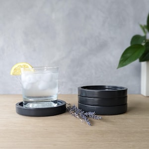 May include: Four black round coasters stacked on top of each other. A glass of water with ice and a lemon wedge sits on a black coaster.