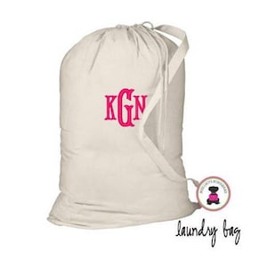 May include: A white canvas laundry bag with a pink monogram "KGN" embroidered on the front. The bag has a drawstring closure and a long handle.