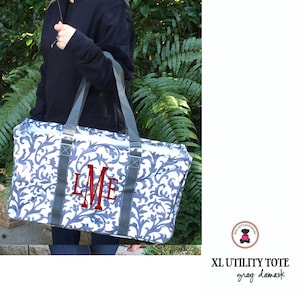 May include: A large gray and white utility tote bag with a damask pattern. The bag has a monogrammed "LME" in red embroidery. The bag is being held by a person.