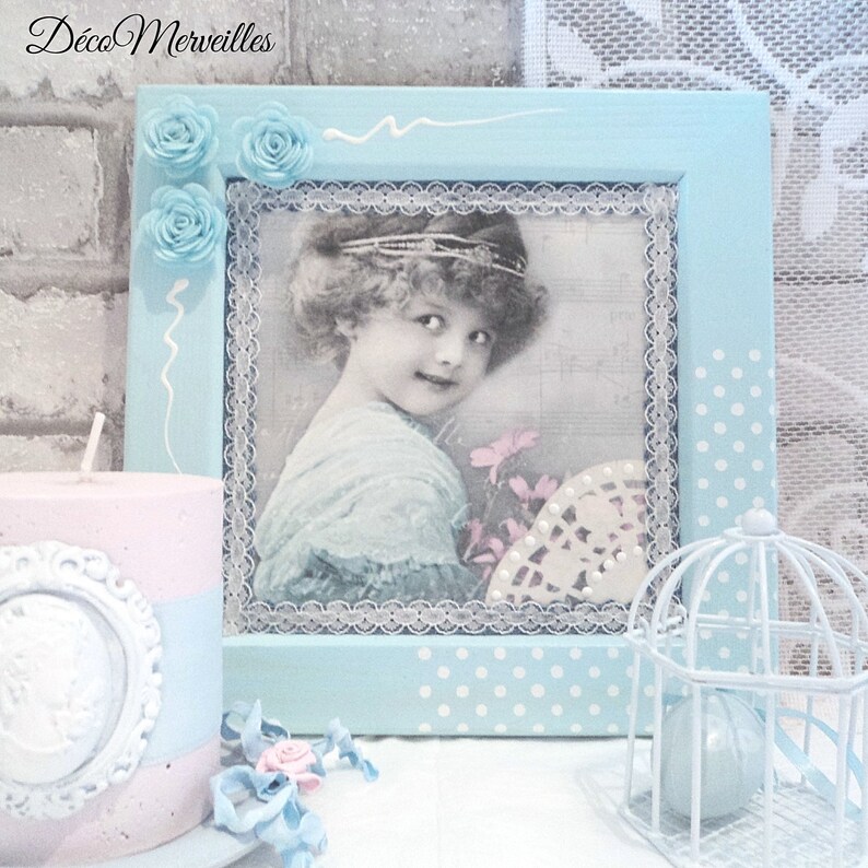 May include: A light blue picture frame with a white lace border and white polka dots. The frame contains a vintage photograph of a young girl with a flower in her hand. The frame is decorated with white swirls and blue flowers.