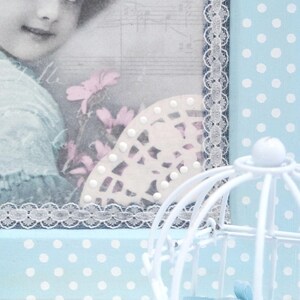 May include: A blue and white polka dot background with a white birdcage in the foreground. A framed picture with a girl and flowers is in the background. The picture has a white lace trim.