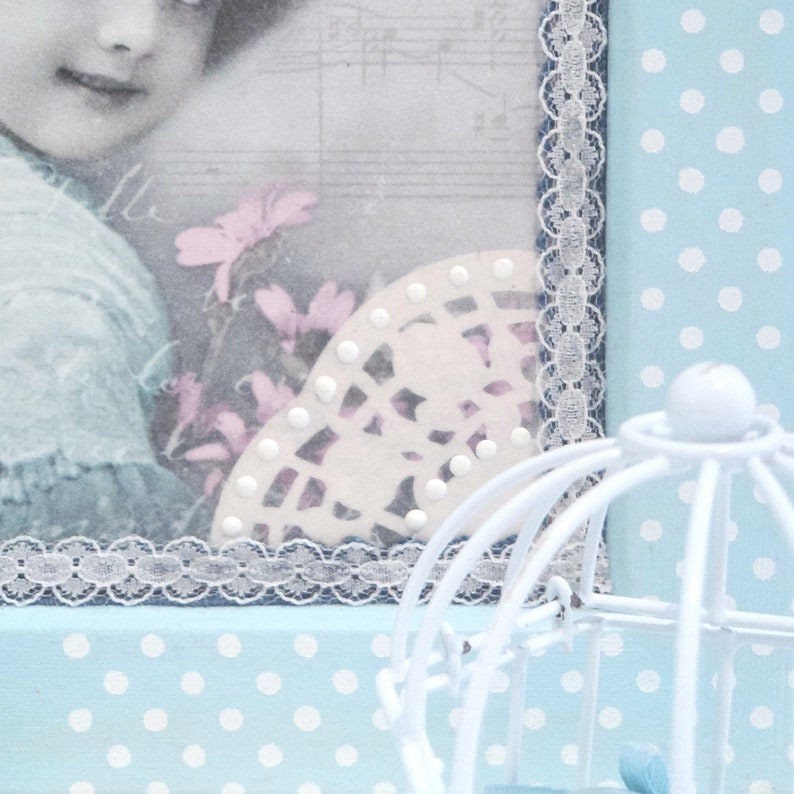 May include: A blue and white polka dot background with a white birdcage in the foreground. A framed picture with a girl and flowers is in the background. The picture has a white lace trim.