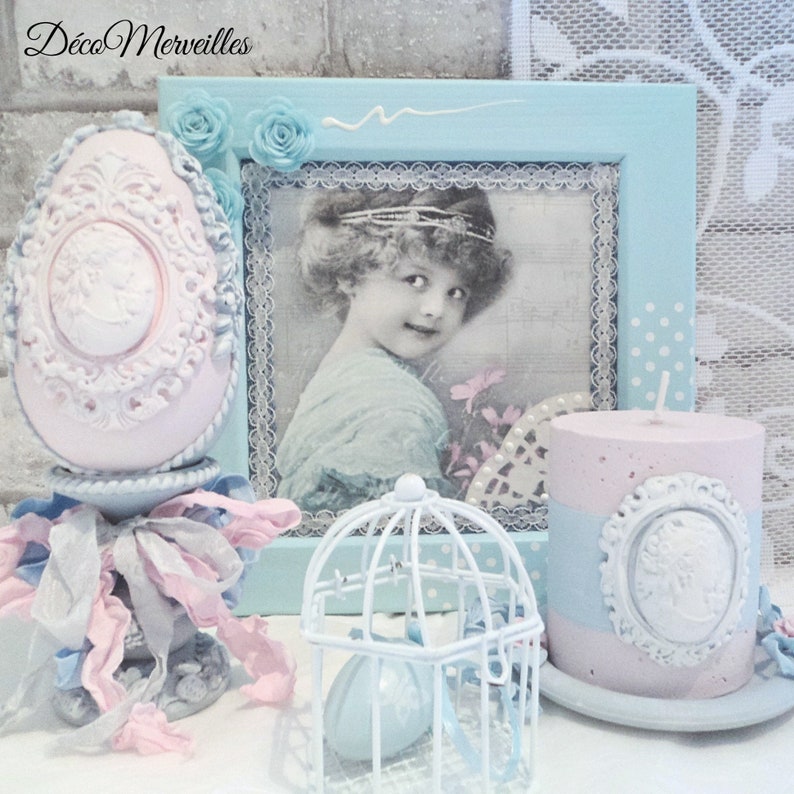 May include: A vintage-inspired decorative set featuring a pink and white egg with intricate details, a blue and white birdcage, and a pink candle with a white cameo design. The set is displayed on a white surface with a blue and white frame in the background.