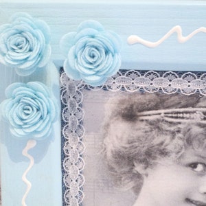May include: A light blue picture frame with white lace trim and three light blue fabric rosettes. The frame holds a black and white photo of a woman wearing a headband.