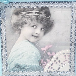 May include: A vintage-style photograph of a young girl with light brown hair wearing a blue dress and a headband. The photo is framed with a white lace border and has a faded, antique look.