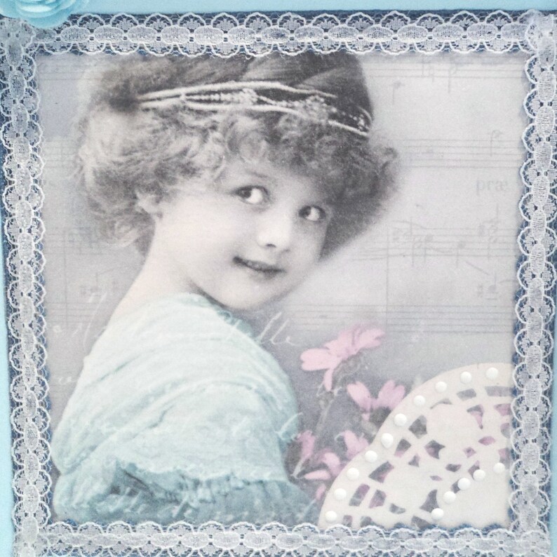 May include: A vintage-style photograph of a young girl with light brown hair wearing a blue dress and a headband. The photo is framed with a white lace border and has a faded, antique look.