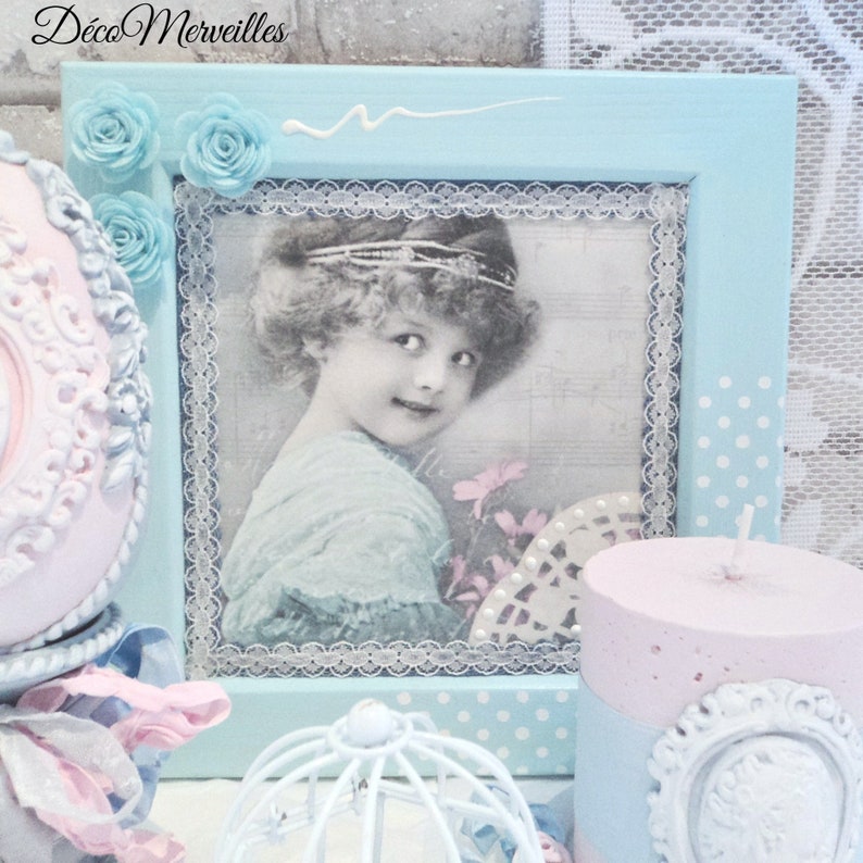 May include: A light blue picture frame with a white lace border and a vintage photo of a young girl wearing a crown. The frame has a polka dot pattern and is decorated with white lace and blue flowers.