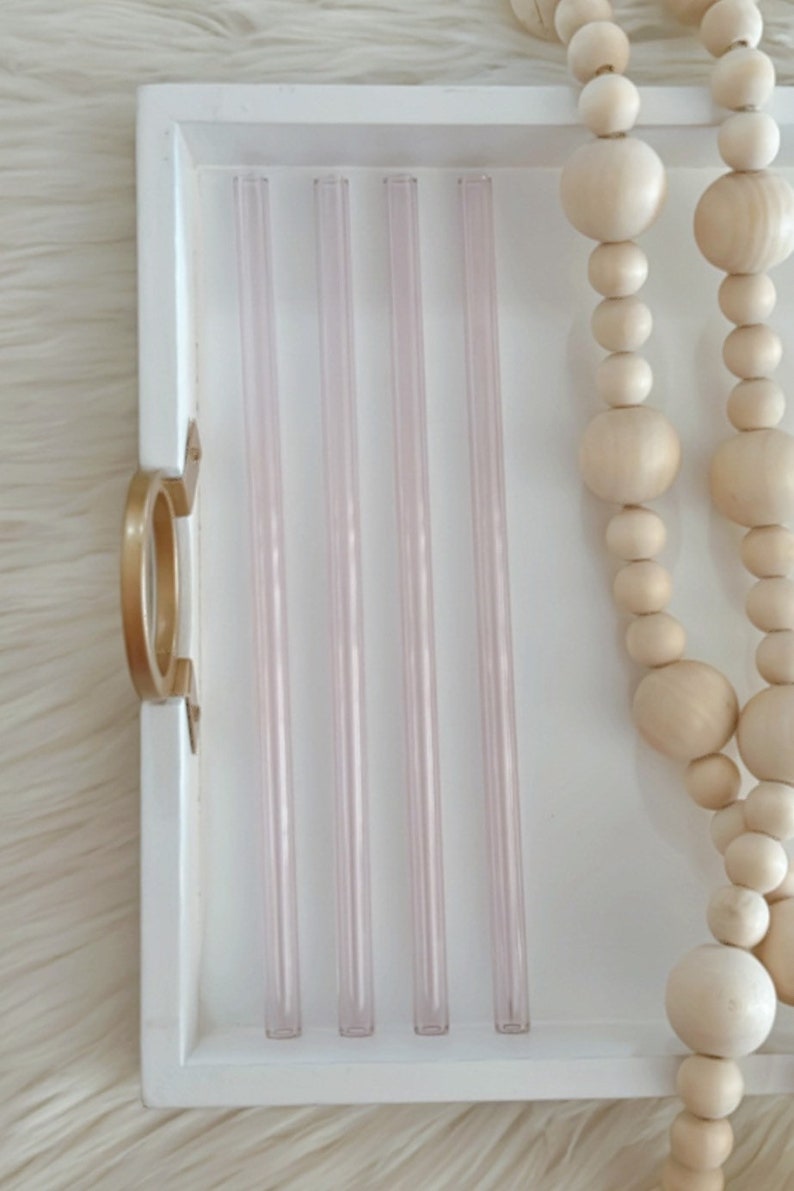 Pu&ograve; includere: Cinque cannucce da bere in vetro trasparente con una leggera sfumatura rosa chiaro sono disposte in fila su un vassoio bianco. Le cannucce sono tutte della stessa lunghezza e larghezza. Il vassoio &egrave; in legno e ha una maniglia dorata.