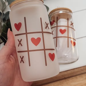 May include: Two glass tumblers with wooden lids. One is frosted and features a tic-tac-toe design with red hearts and brown X's. The other is clear with the same design. Both tumblers are designed for cold beverages.