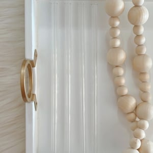 Pu&ograve; includere: Un vassoio bianco con una maniglia dorata contiene sei cannucce di vetro trasparente. Una collana di perline di legno di colore naturale pende dal bordo del vassoio.