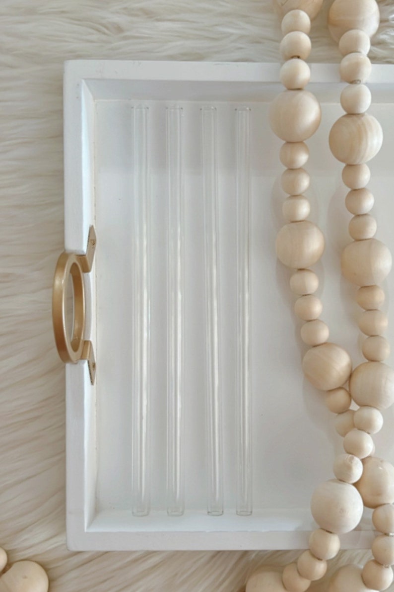 Pu&ograve; includere: Un vassoio bianco con una maniglia dorata contiene sei cannucce di vetro trasparente. Una collana di perline di legno di colore naturale pende dal bordo del vassoio.