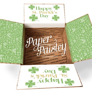 May include: Open cardboard box with a green shamrock pattern on the inside and a white lid with the text "Happy St. Patrick's Day" in gold lettering. The box is decorated with green shamrocks and the text "Paper Paisley" is written in white on the side of the box.