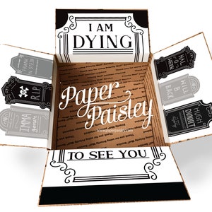 May include: A brown cardboard box with a black and white design. The box is open and the inside is lined with brown paper. The box has a black and white design with the words "I am Dying to See You" on the front. The box is decorated with black and white tombstone-shaped stickers with various names and phrases.