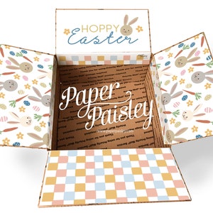 May include: A cardboard box with a checkered pattern on the bottom and a bunny rabbit pattern on the sides. The box is open and the top flap has the text "HOPPY Easter" with a bunny rabbit illustration. The text "Paper Paisley" is written on the inside of the box.