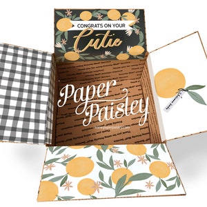 May include: Open cardboard box with a black and white checkered pattern on the inside. The box is decorated with a lemon and floral pattern. The top of the box has a black background with white text that reads "Congrats on your Cutie".