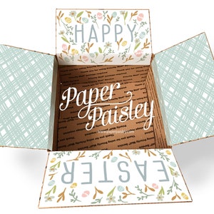 Puede incluir: Una caja de cartón abierta con un diseño floral en las solapas superior e inferior. La caja está decorada con las palabras "Feliz Pascua" y "Paper Paisley".