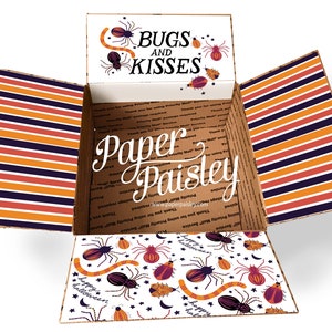 May include: An open cardboard box with a Halloween theme. The box is lined with white paper featuring a pattern of spiders, worms, and stars. The top of the box has a white label with the text "BUGS AND KISSES" and a spider design. The box is labelled "Paper Paisley".