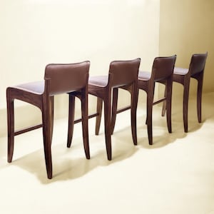 May include: Four brown leather bar stools with dark wood frames. The stools are arranged in a row, with the backs of the stools facing the camera.