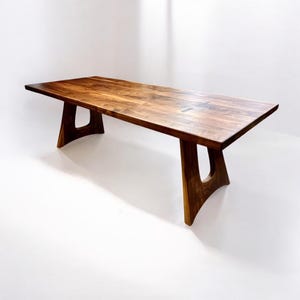 May include: A long, rectangular wooden dining table with a dark brown finish. The table has a unique, modern design with two large, curved legs on each side. The legs are made of the same wood as the table top and have a cutout design.